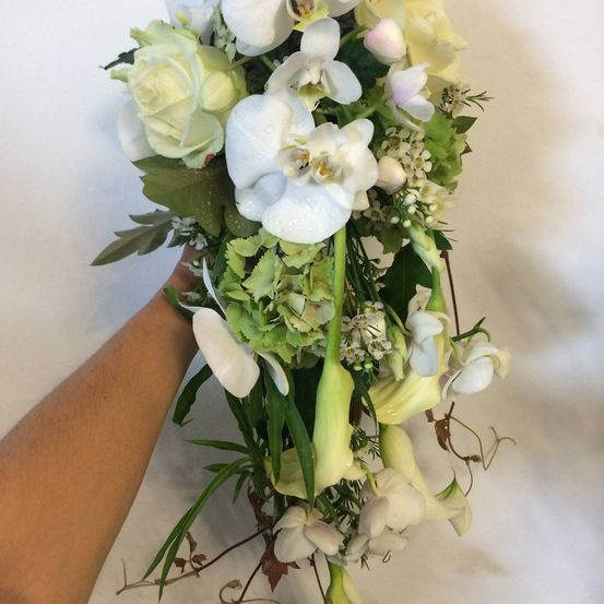 Ein Strauß weißer Blumen, einschließlich Orchideen, Rosen und Lilien, von einer Hand gehalten vor weißem Hintergrund.