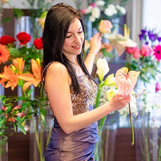 Eine Frau in einem lila Kleid hält eine rosa Blume vor einer bunten Blumenarrangement.