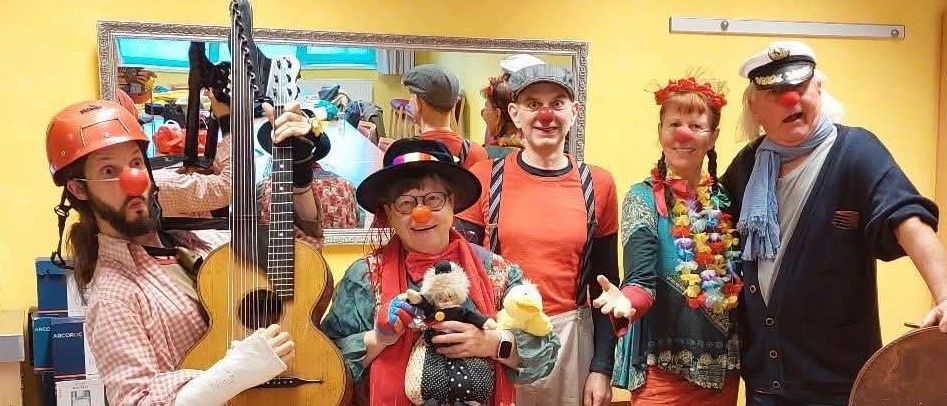 Eine Gruppe von Menschen, die als Clowns verkleidet sind, posiert für ein Foto in einem leuchtend farbigen Raum mit Gitarren, Plüschtieren und Hüten.