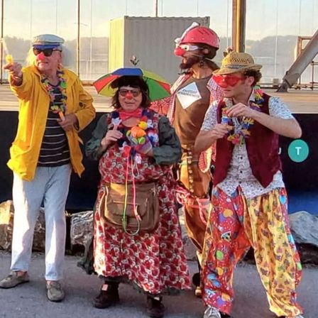 Vier Menschen in bunten Clownkostümen stehen zusammen auf einer Bühne, zwei halten Requisiten und einer ein Mikrofon. Im Hintergrund befindet sich eine Glaswand und eine Struktur.