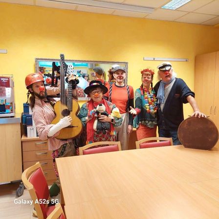 Fünf Menschen in Clownkostümen posieren für ein Foto in einem Raum. Jeder hält Requisiten, darunter eine Gitarre, ein Schlagzeug und Spielzeug. Ein Tisch und Stühle sind im Vordergrund.