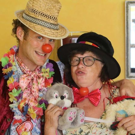 Ein Mann mit Strohhut und Clownsnase lächelt neben einer Frau mit schwarzer Mütze, Brille und rotem Schleifenband. Beide halten Plüschtiere und stehen vor einer gelben Wand.