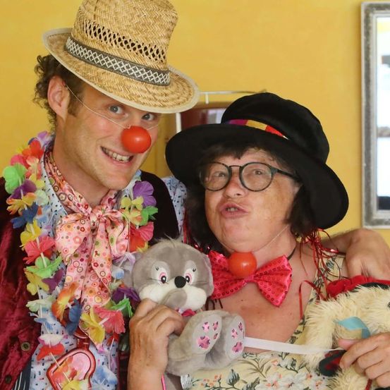 Ein Mann mit Strohhut und Clownsnase lächelt neben einer Frau mit schwarzer Mütze, Brille und rotem Schleifenband. Beide halten Plüschtiere und stehen vor einer gelben Wand.