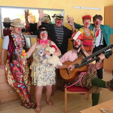 Eine Gruppe von Clowns in bunten Kostümen posiert für ein Foto in einem Raum, wobei einer Gitarre spielt und ein anderer Kuscheltiere hält.