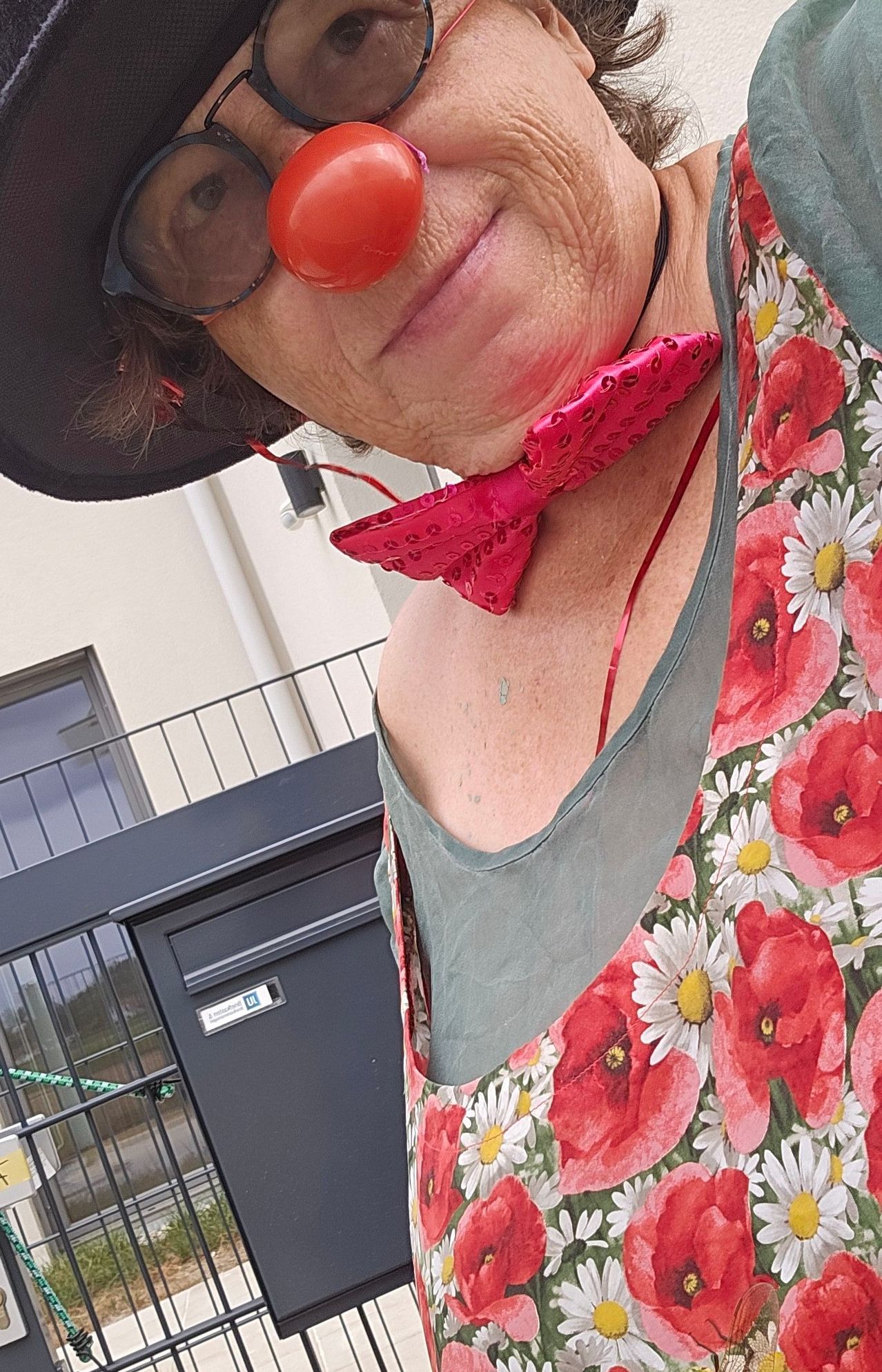 Eine Frau trägt ein Blumenkleid mit roten Mohnblumen und eine rosa Fliege um den Hals, vor einem Gebäude mit einem Briefkasten stehend.
