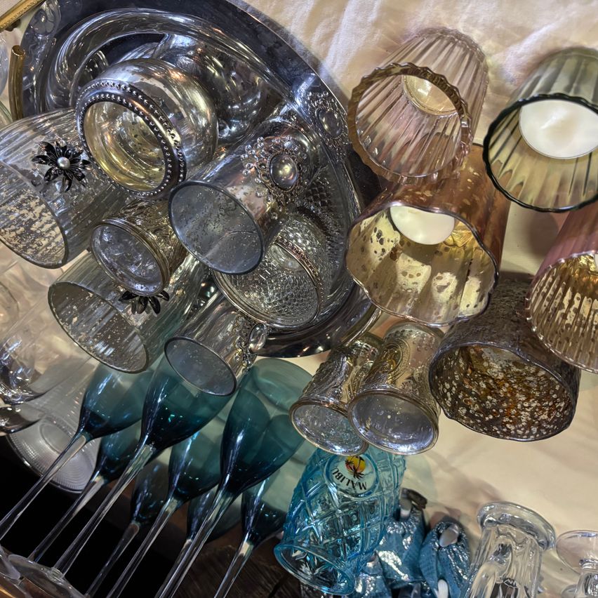 Eine Sammlung eleganter Gläser auf einem weißen Tuch umfasst silberne und goldgerandete Gläser, einige mit dekorativen Elementen und Kerzen, neben hohen blauen Champagnergläsern und einer blauen Flasche.