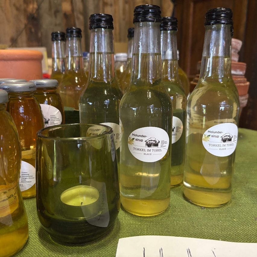 Mehrere Flaschen mit Honigsirupetiketten sind auf einem grünen Tisch ausgestellt. Ein Glas mit einer brennenden Kerze steht davor.