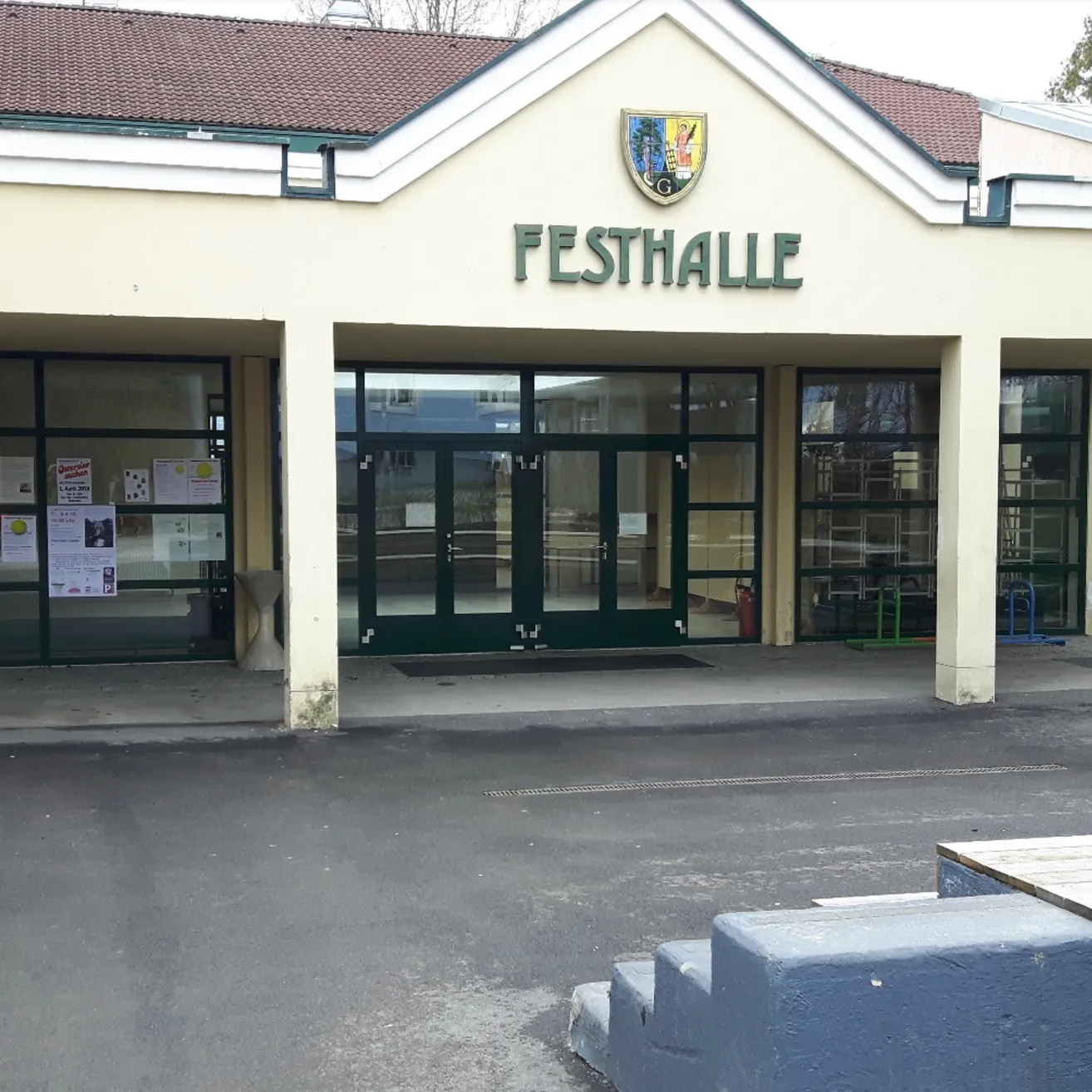 Der Vordereingang eines Gebäudes namens 'Festhalle' mit Glastüren und Fenstern, einem Logo über dem Eingang und einem Parkplatzschild rechts.