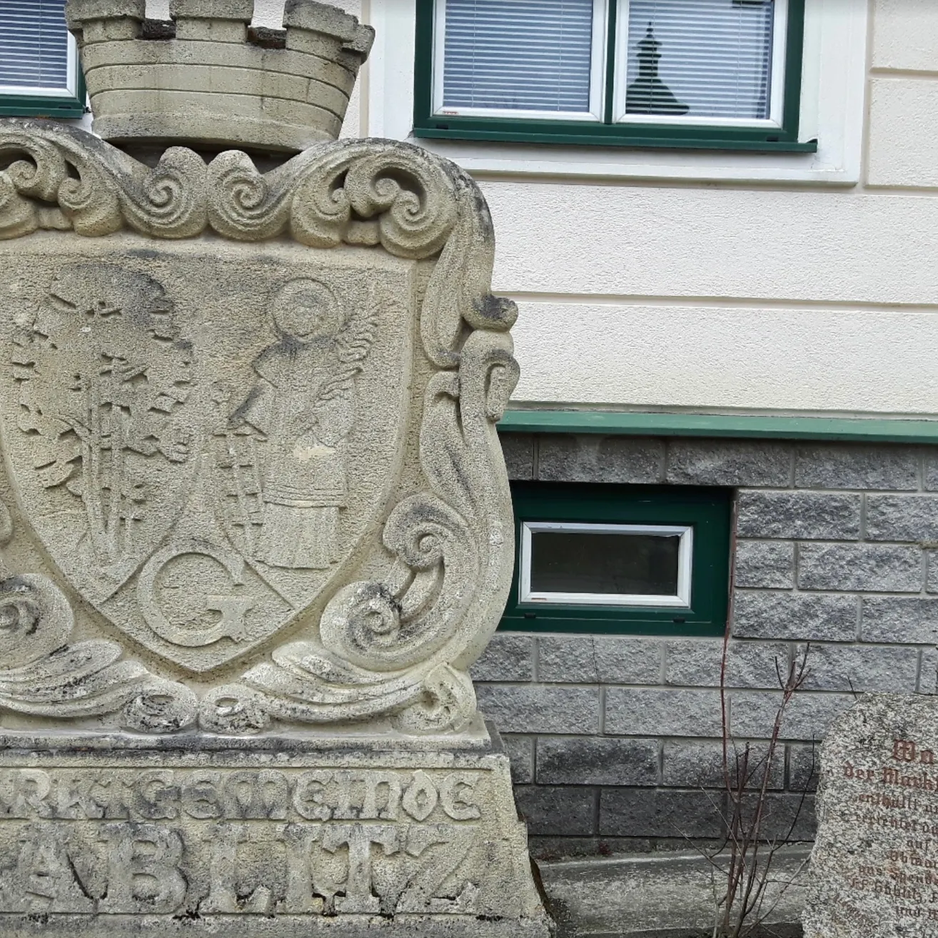 Ein Steinmonument mit einem Wappen und einer Krone auf der Spitze, vor einem Gebäude mit grünen Fenstern.