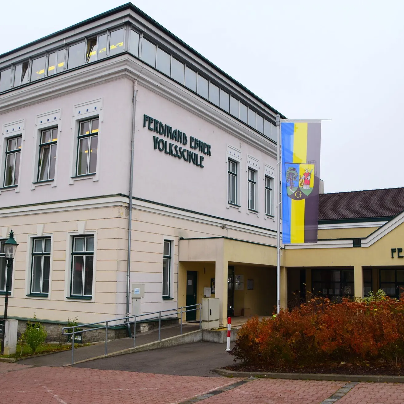 Ein weißes Gebäude mit vielen Fenstern und einem Fahnenmast vor dem Gebäude. Das Gebäude hat ein Namensschild mit der Aufschrift 'FERDINAND EINWER VOLKSSCHULE'.