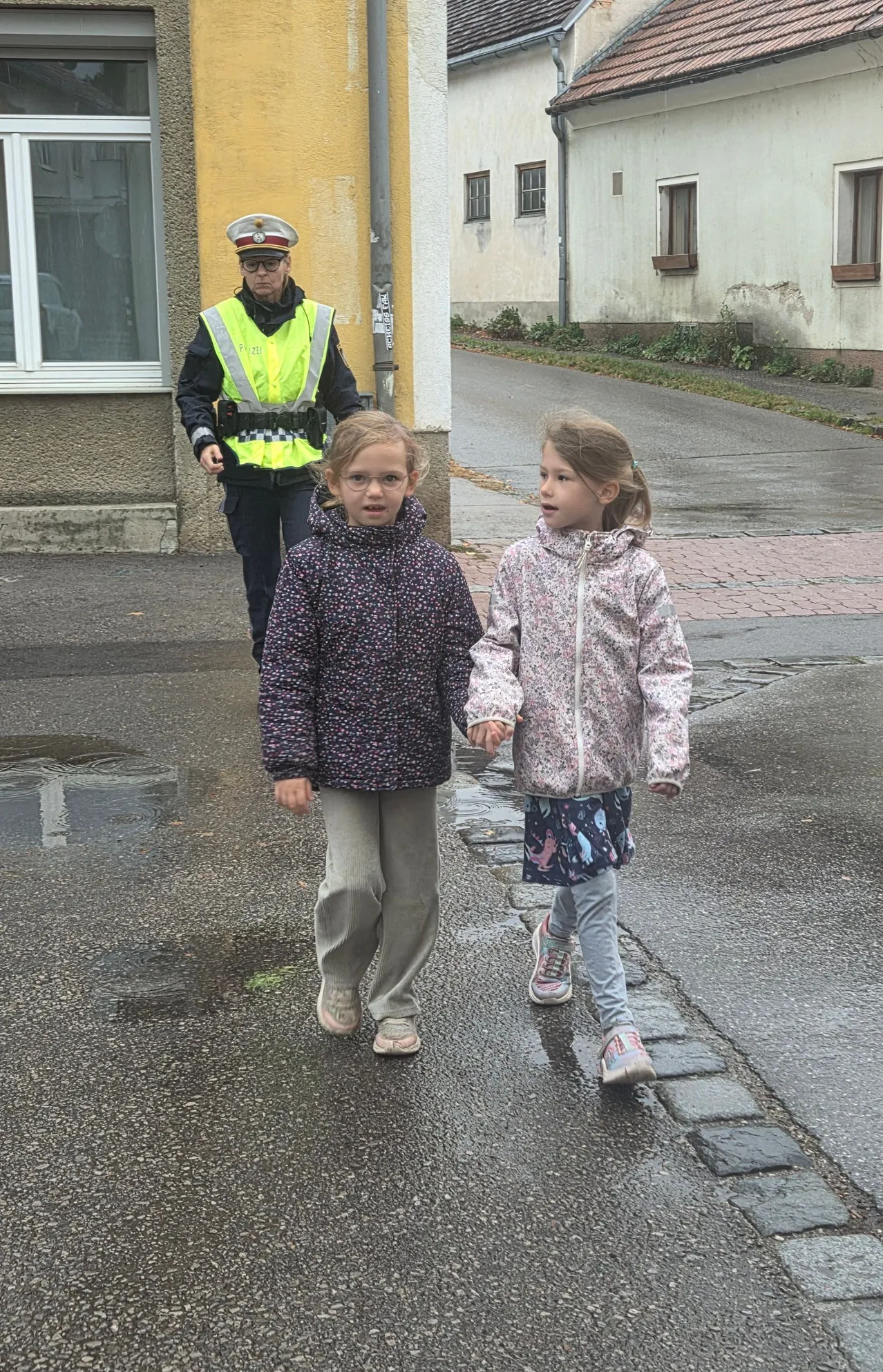 Zwei junge Mädchen gehen Hand in Hand auf einer nassen Straße. Dahinter läuft eine Polizistin in einem reflektierenden Weste.