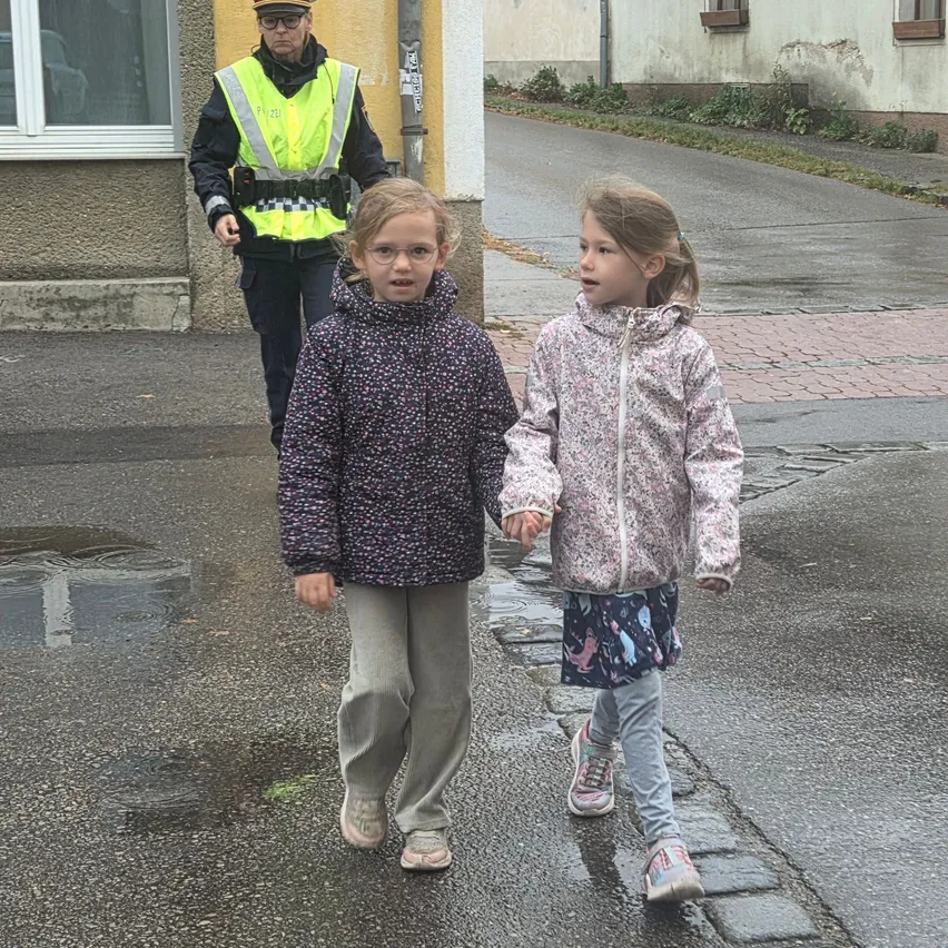Zwei junge Mädchen gehen Hand in Hand auf einer nassen Straße. Dahinter läuft eine Polizistin in einem reflektierenden Weste.