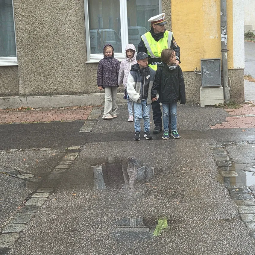 Ein Verkehrspolizist führt eine Gruppe von Kindern über eine nasse Straße vor einem gelben Gebäude.