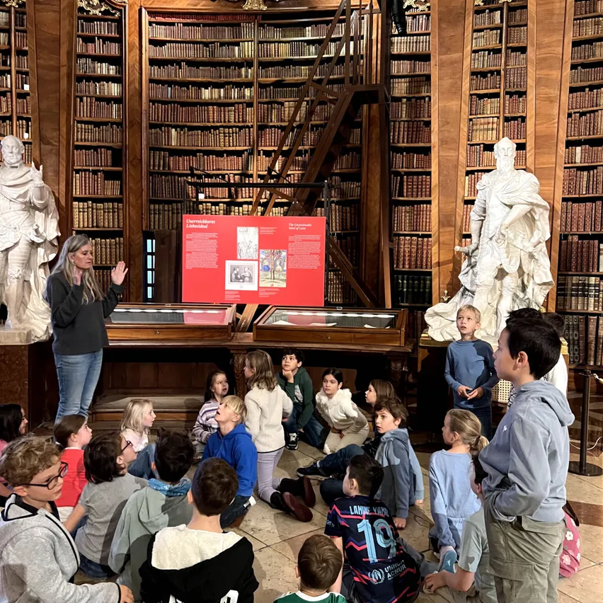 Eine Frau hält eine Präsentation für eine Gruppe von Kindern in einer Bibliothek. Die Kinder sitzen auf dem Boden, und einige stehen. Auf dem Tisch vor ihnen befindet sich ein roter Plakat.