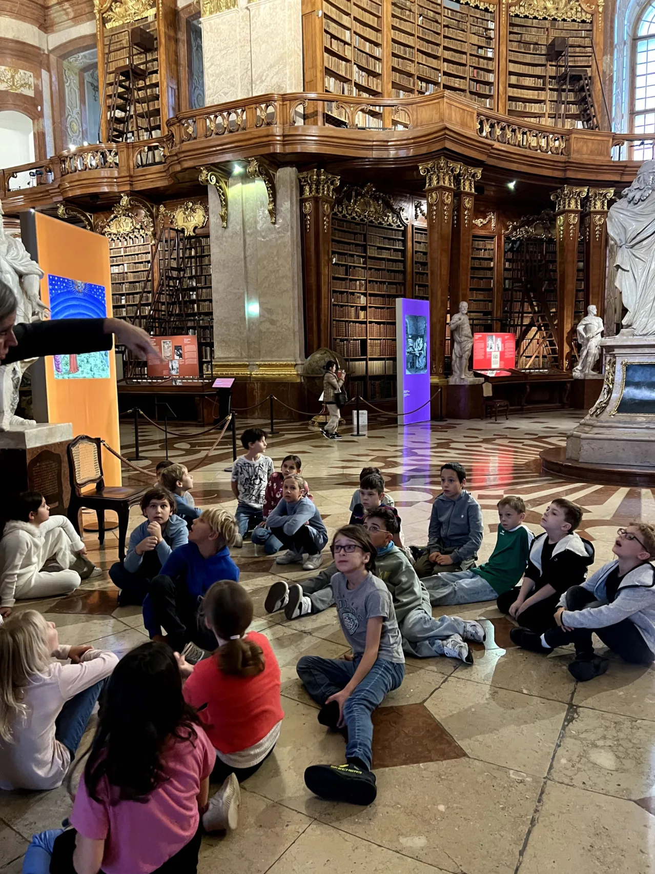 Eine Gruppe von Kindern sitzt auf dem Boden in einer Bibliothek und hört einer Frau zu, die auf einen Bildschirm zeigt.