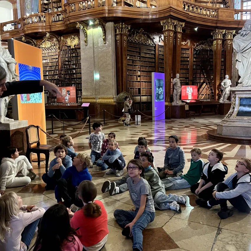 Eine Gruppe von Kindern sitzt auf dem Boden in einer Bibliothek und hört einer Frau zu, die auf einen Bildschirm zeigt.