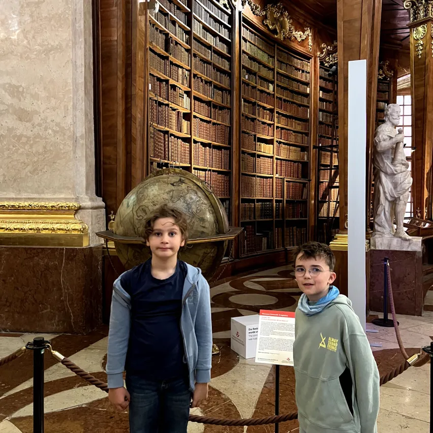 Zwei junge Jungen stehen in einer Bibliothek mit einer großen Globus im Vordergrund und einer Statue hinter ihnen.