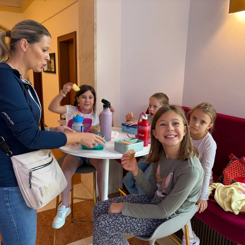Eine Frau in einer blauen Jacke steht vor einem Tisch, an dem vier Mädchen sitzen. Sie essen Snacks und trinken aus Flaschen.