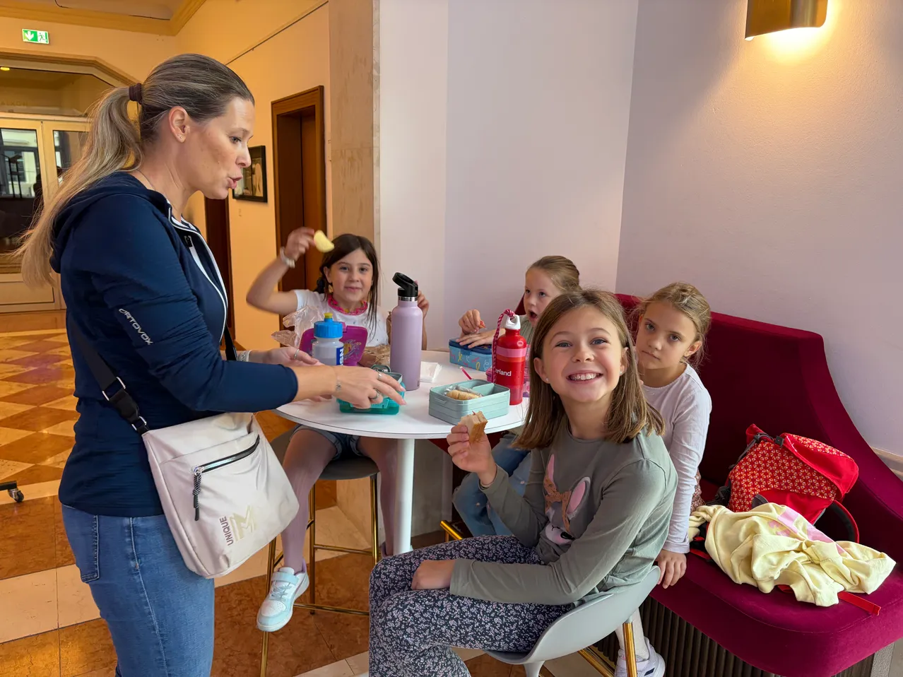 Eine Frau in einer blauen Jacke steht vor einem Tisch, an dem vier Mädchen sitzen. Sie essen Snacks und trinken aus Flaschen.