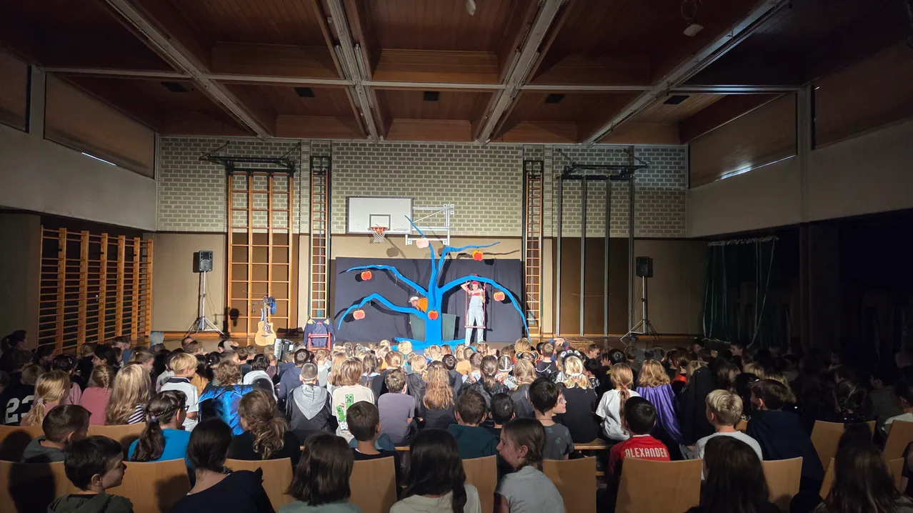 Eine Gruppe von Menschen sieht eine Aufführung auf einer Bühne mit einem blauen Baumhintergrund.