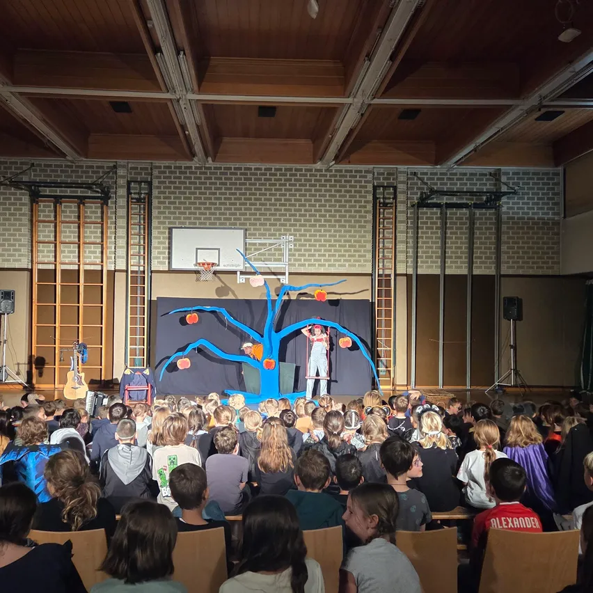 Eine Gruppe von Menschen sieht eine Aufführung auf einer Bühne mit einem blauen Baumhintergrund.