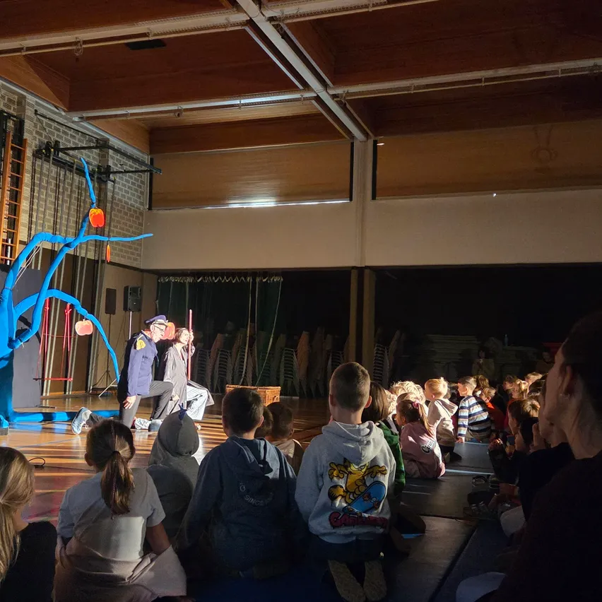 Eine Gruppe von Kindern und Erwachsenen schaut sich eine Aufführung auf einer Bühne in einer Turnhalle an. Die Bühne hat eine blaue Baumattrappe und eine Person spielt Akkordeon.