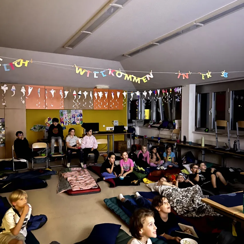 Eine Gruppe von Kindern sitzt auf dem Boden in einem Klassenzimmer. Sie tragen Freizeitkleidung und einige halten Teller mit Essen.