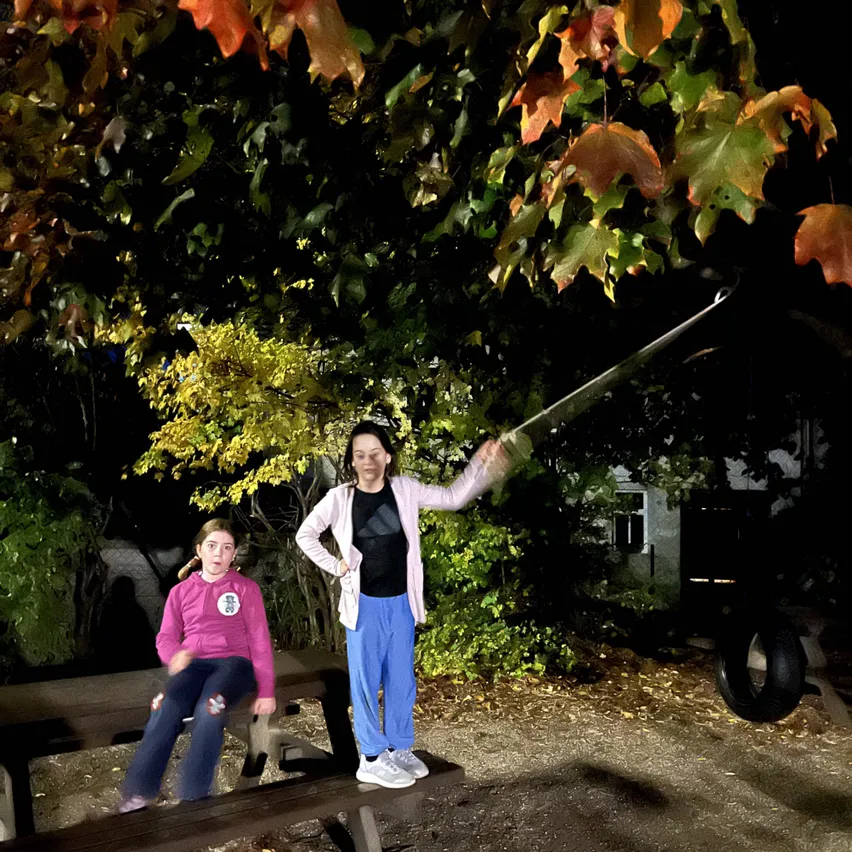 Zwei Frauen sitzen bei Nacht auf einer Bank im Park. Eine von ihnen hält ein Schwert.