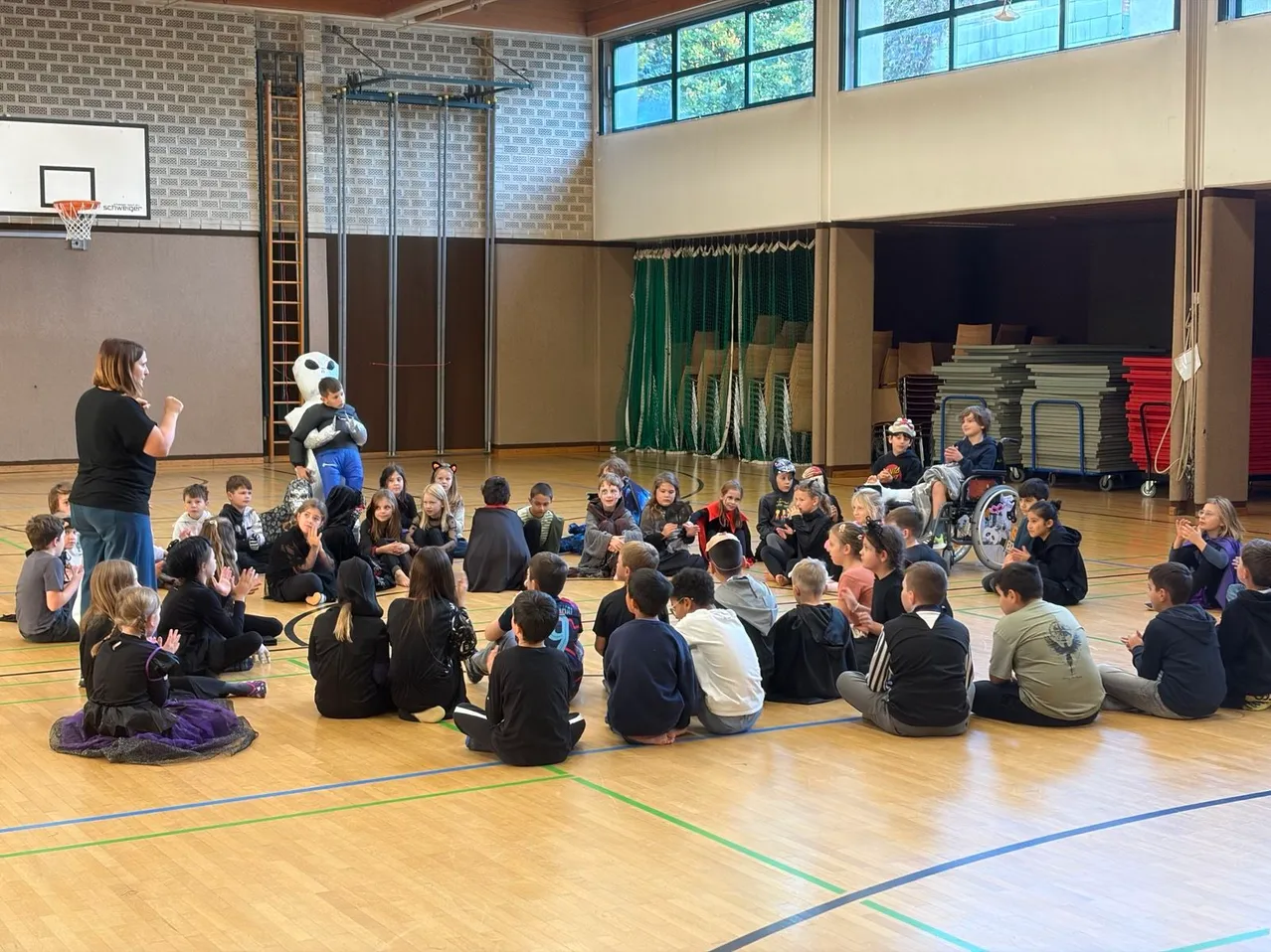 Eine Gruppe von Kindern und einer Frau sitzen im Kreis auf einem Basketballplatz. Ein Maskottchen steht hinter der Frau.