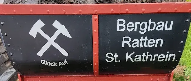 Ein roter und schwarzer Kohlewagen auf einer Holzschiene mit einem Haufen Kohle oben drauf. Es steht Bergbau Ratten St. Kathrein.