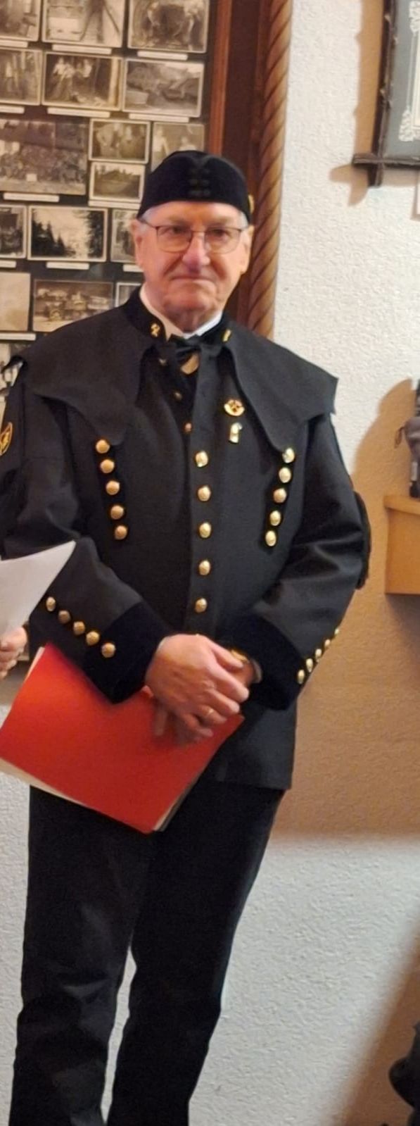 Ein Mann in einem schwarzen Uniform mit goldenen Knöpfen und einer Fliege hält eine rote Mappe in den Händen.