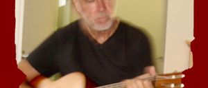 Ein älterer Mann spielt drinnen eine akustische Gitarre und konzentriert sich darauf. Er trägt ein schwarzes T-Shirt und graues Haar.