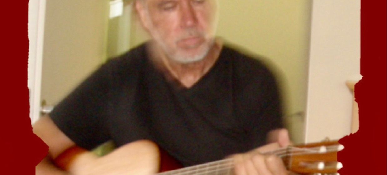 Ein älterer Mann spielt drinnen eine akustische Gitarre und konzentriert sich darauf. Er trägt ein schwarzes T-Shirt und graues Haar.