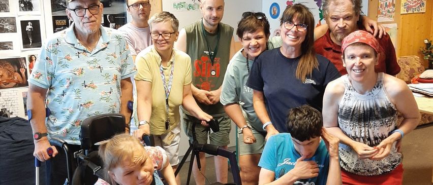 Eine Gruppe von Menschen, darunter zwei Kinder im Rollstuhl, posiert für ein Foto in einem Raum mit einer weißen Wand und verschiedenen Bildern.