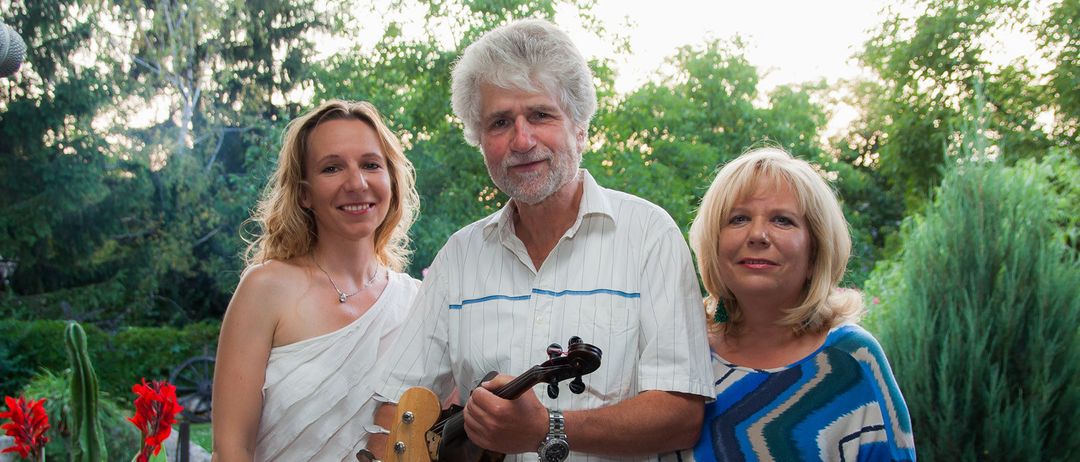 Ein Mann und zwei Frauen posieren für ein Foto im Garten. Der Mann hält eine Geige und eine Gitarre. Die Frau links trägt ein weißes Kleid. Die Frau rechts trägt ein blau-weiß gemustertes Oberteil.