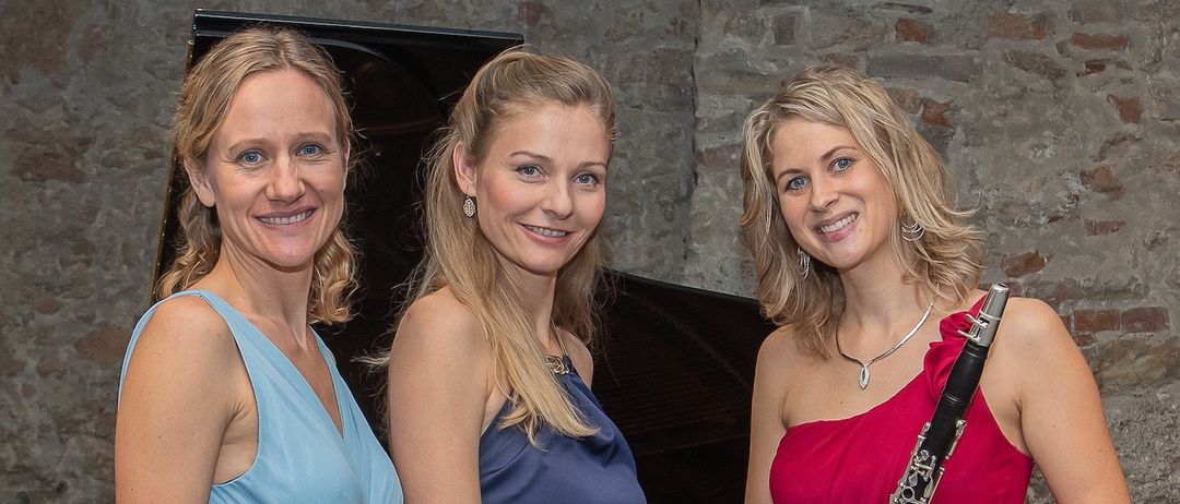 Drei Frauen in eleganten Kleidern, lächelnd und posieren für ein Foto mit einem Klavier im Hintergrund. Eine hält eine Klarinette, eine andere eine Halskette.