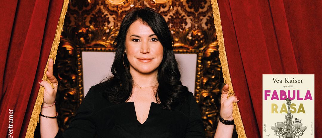 Eine Frau steht mit erhobenen Händen neben einem roten Vorhang und trägt ein schwarzes Kleid. Hinter ihr befindet sich eine goldfarbene Wand und ein Kronleuchter. Ein Buch mit dem Titel 'FA' ist teilweise rechts sichtbar.