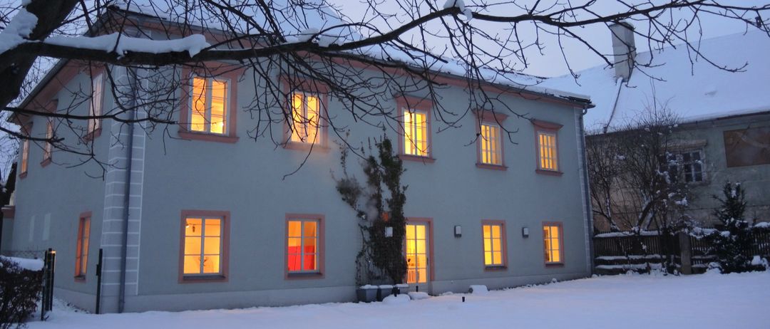 Ein großes weißes Haus mit rosa Umrandung und mehreren von warmem Licht erleuchteten Fenstern steht in einer verschneiten Landschaft.