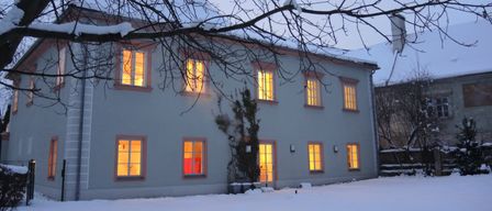 Ein großes weißes Haus mit rosa Umrandung und mehreren von warmem Licht erleuchteten Fenstern steht in einer verschneiten Landschaft.