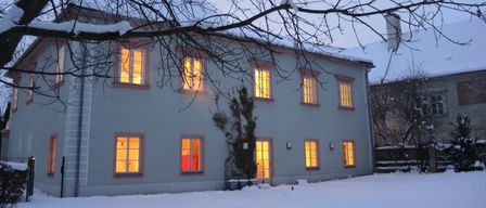 Ein weißes Haus mit schneebedecktem Dach und leuchtenden Fenstern im Winter. Die Vorderseite ist schneebedeckt.