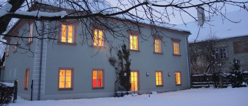 Ein weißes Haus mit schneebedecktem Dach und leuchtenden Fenstern im Winter. Die Vorderseite ist schneebedeckt.