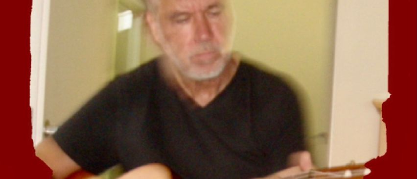 A man with a beard is playing a guitar. He is wearing a black t-shirt. The guitar has a wooden body and metal frets.