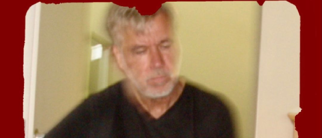 Ein älterer Mann spielt Gitarre in einem schwach beleuchteten Raum. Er trägt ein schwarzes T-Shirt und hat einen konzentrierten Gesichtsausdruck. Die Gitarre hat einen braunen Korpus und eine weiße Schlagplatte.