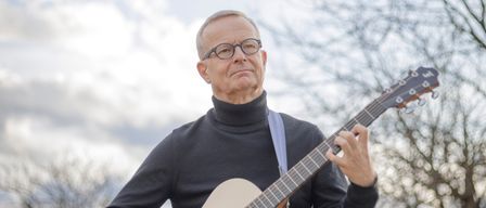 Ein Mann in einem schwarzen Rollkragenpullover spielt draußen eine Gitarre. Die Gitarre hat einen braunen Korpus und schwarze Besaitung. Der Himmel ist blau mit weißen Wolken.