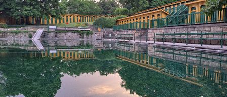 Ein ruhiger Freibad mit einem gelben und grünen Gebäude an der Seite, das sich auf der Wasseroberfläche spiegelt. Bäume und Pflanzen umgeben das Gebiet.