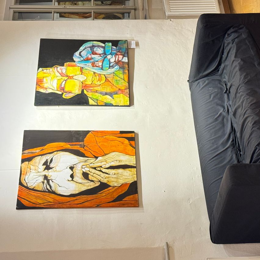 Zwei abstrakte Gemälde hängen an einer weißen Wand. Das obere Gemälde zeigt eine leuchtende, mehrfarbige Figur mit einer Maske. Das untere Gemälde zeigt eine Figur mit einem ruhigen Gesicht, den Händen an den Wangen und orangefarbener Kleidung. Ein schwarzer Sessel befindet sich rechts von den Gemälden.