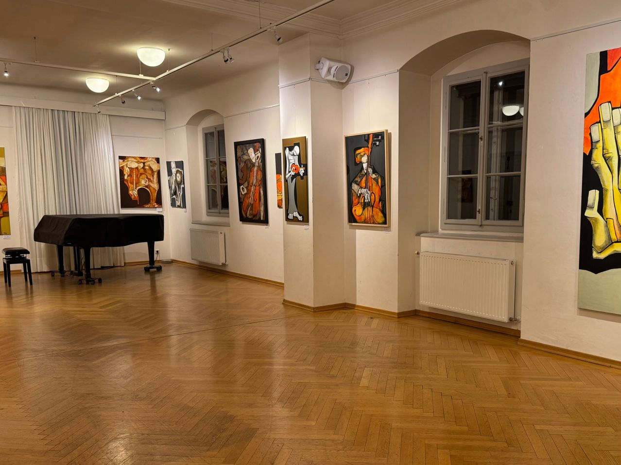 Ein leerer Raum mit Holzböden und weißen Wänden, mit mehreren Gemälden an den Wänden, einem Klavier in der Nähe der Wand und einem Heizkörper unter dem Fenster.