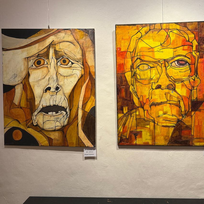 Zwei abstrakte Gesichtsgemälde hängen an einer weißen Wand. Das linke Gemälde zeigt eine Person mit langem Bart und Hut. Das rechte Gemälde zeigt eine Person mit Brille und Schnurrbart.