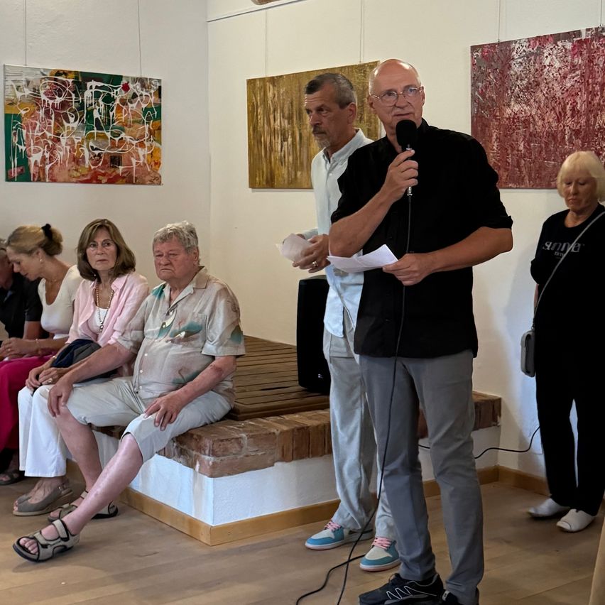 Ein Mann spricht in ein Mikrofon vor einem Publikum in einer Kunstgalerie, mit abstrakten Gemälden an der Wand.