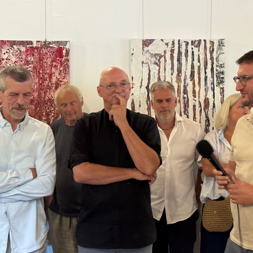 Eine Gruppe von Männern und Frauen steht vor einer Wand mit zwei abstrakten Gemälden. Ein Mann hält ein Mikrofon und spricht, während die anderen zuhören.